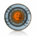 A round ceramic plate featuring a central sun motif with a stylized face and radiating gold lines, surrounded by concentric rings of dark gray and light blue, finished with a thin gold rim.