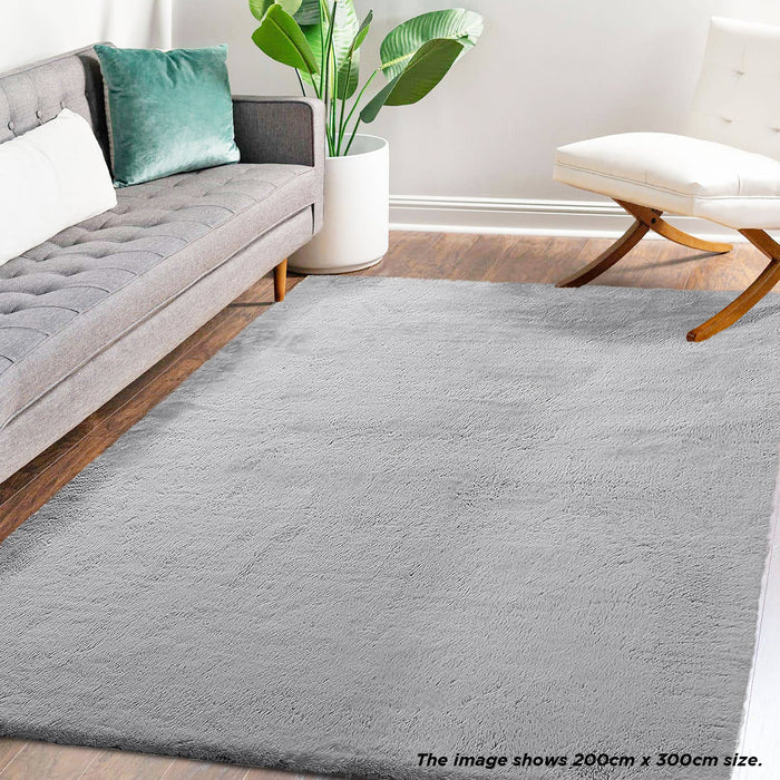 Azaliya 200x300cm Faux Rabbit Fur Floor Rug in Silver, styled in a modern living room with a gray mid-century sofa, green pillow, white planter, and wooden-legged lounge chair.