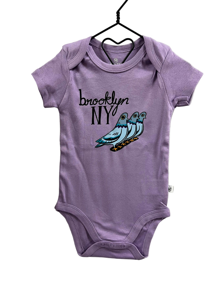 A lavender baby bodysuit hanging on a black hanger against a light gray wall. The bodysuit features a graphic print of three blue and teal pigeons with the text 'brooklyn NY' in black script and block lettering.