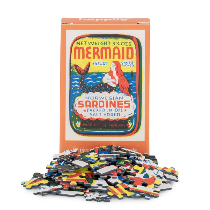 A box with Louise Lockhart’s retro sardines design labeled “Mermaid | 99 Piece Mini Jigsaw Puzzle” from Happily Jigsaw Puzzles stands behind a pile of loose puzzle pieces from this charming mini puzzle.